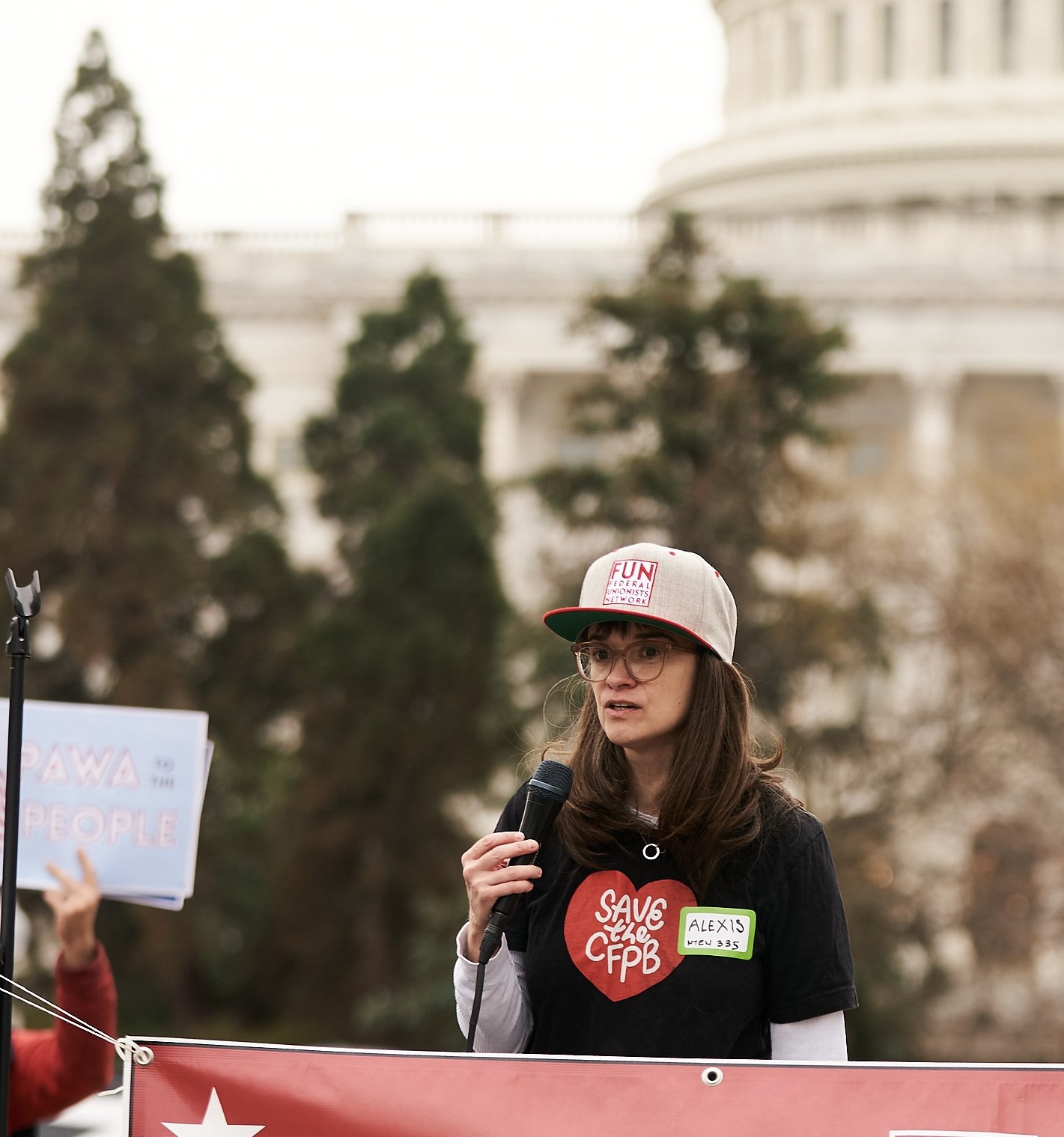 A photo of Alexis Goldstein in front of the U.S. Capitol building, wearing a black shirt with a red heart that reads 'Save the CFPB' inside the heart. She is holding a microphone in her hand, and to her left is a sign that reads 'PAWA to the People'